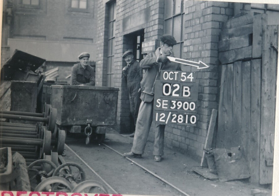 Three men stand outside a brick industrial building; one holds a sign reading “OCT 54 02B SE 3900 12/2810” with an arrow, while mining carts and machinery are visible on rails nearby.